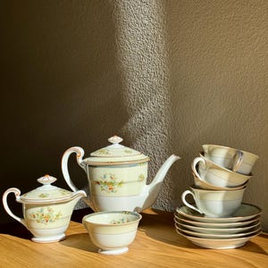 Może przedstawiać: Kremowy, zabytkowy zestaw do herbaty z czajnikiem, śmietanką, cukiernicą, filiżankami i spodkami. Zestaw ma kremową podstawę z kwiatowymi i złotymi detalami. Czajnik ma zakrzywioną wylewkę i uchwyt. Filiżanki są ułożone w stos.