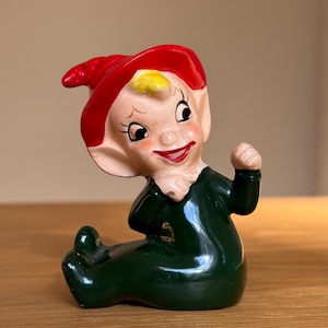 May include: A ceramic elf figurine, seated and wearing a red hat and green outfit. The elf has a smiling face with rosy cheeks and a yellow tuft of hair. The figurine is posed with one fist clenched, a vintage decorative item.