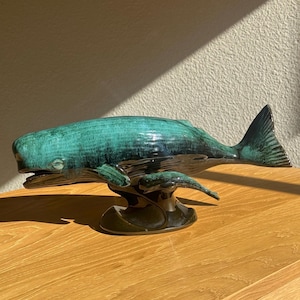 Op de afbeelding: Een keramieken walvisbeeldje met een turquoise en donkergroene glazuur. De walvis is gemonteerd op een donkergroene voet en staat op een houten oppervlak. Het beeldje heeft een gedetailleerd ontwerp en is een decoratief item.