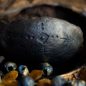 Op de afbeelding: Een donkergrijze, handgesneden stenen kom met geëtste geometrische ontwerpen. De kom heeft een ruwe textuur en is omgeven door kleine, metallic blauwe kralen en gedroogde bladeren, wat een rustieke, aardse esthetiek creëert.