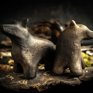 Puede incluir: Dos figuras de oso hechas a mano en tonos marrón oscuro y gris. Un oso está de pie, el otro está sentado. Las esculturas están colocadas sobre una superficie oscura y texturizada, con un fondo borroso.