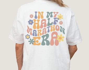 Op de afbeelding: Wit T-shirt met een retro-grafische afbeelding met de tekst "In My Half Marathon Era" met kleurrijke bloemen en smiley-gezichten.