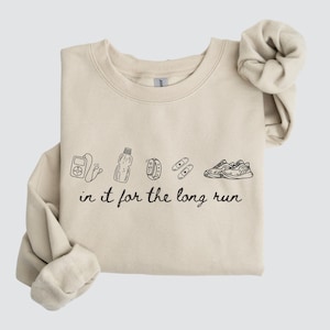 May include: Sand-colored sweatshirt with black line art illustrations of medical devices, a water bottle, a smartwatch, bandages, and running shoes. The phrase "in it for the long run" is printed below the illustrations. The sweatshirt has a crew neck and long sleeves.