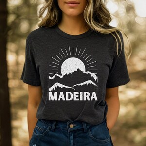 Puede incluir: Una camiseta gris oscuro con un gráfico blanco de un sol poniéndose detrás de las montañas y la palabra "MADEIRA" impresa debajo.