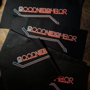 May include: Several black fabric patches with the word "GOODNEIGHBOR" in red and light blue lettering. The text is accompanied by a stylized arrow design. The patches are arranged on a wooden surface.