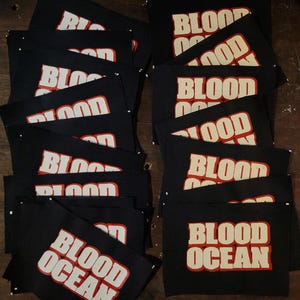 Pode incluir: Uma coleção de patches de tecido preto com as palavras "BLOOD OCEAN" impressas em uma fonte arrojada vermelha e branca. Os patches retangulares são dispostos em um padrão ligeiramente sobreposto, sugerindo um pedido em massa ou exibição.