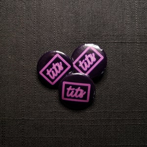 May include: Three round, dark purple buttons with the word "tits" in a pink neon-style font inside a pink square or rectangle. The buttons are arranged on a dark, textured surface.
