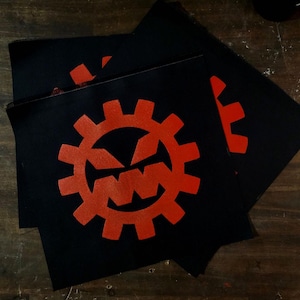 May include: Black fabric patches with a red gear design featuring a face. The patches are square-shaped and stacked on a wooden surface. The gear design has a face in the center.