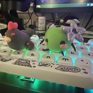 May include: Two small, handmade clay figurines. One is a black and red chicken with a pink bow. The other is a green apple with a purple flower on top. The figurines are sitting on a white keyboard with black keys.