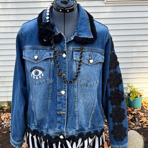 May include: A blue denim jacket with a black faux fur collar, black floral embroidery on the sleeves, and a black and white striped ruffle at the bottom. The jacket has a large eye patch on the front and a pearl necklace.