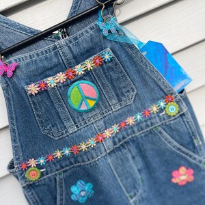 以下が含まれることがあります： カラフルな刺繍の花と平和のサインのパッチが付いた青いデニムのオーバーオール。オーバーオールは、ボタン留めの小さなポケットが前面にあります。