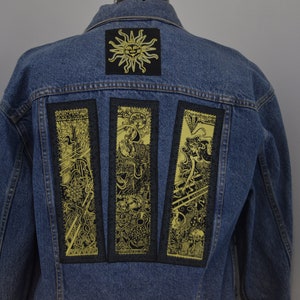 May include: A blue denim jacket with three yellow and black patches on the back. The patches feature intricate designs with a sun motif.