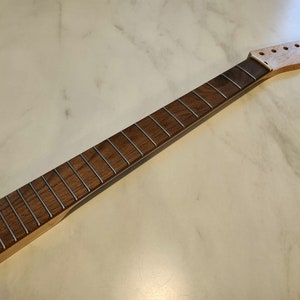 Può includere: Un manico di chitarra con tastiera marrone scuro e tasti argentati. La paletta è in legno chiaro e presenta fori preforati per le meccaniche. Il manico è incompiuto, pronto per la personalizzazione. Un pezzo di strumento musicale.