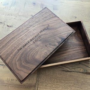 Könnte beinhalten: Holzkiste mit Scharnierdeckel, teilweise geöffnet, wodurch das Innere sichtbar wird. Der Deckel ist mit dem Text "Five years in and I would still pick you! 12.05.2020" graviert. Die Kiste ist aus dunklem Holz mit sichtbarer Maserung gefertigt.