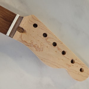 May include: Close-up of a guitar neck and headstock. The neck is dark brown, and the headstock is light-colored wood with six round holes for tuning pegs. The headstock has a curved shape.