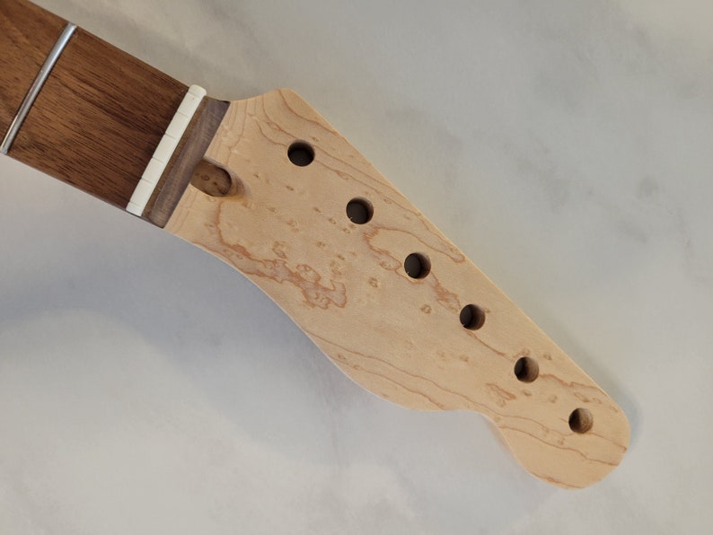 May include: Close-up of a guitar neck and headstock. The neck is dark brown, and the headstock is light-colored wood with six round holes for tuning pegs. The headstock has a curved shape.