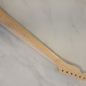May include: A light-colored, unfinished guitar neck with six tuning peg holes. The wood grain is visible, and the neck is angled diagonally against a light-colored background. This guitar part is ready for customization.