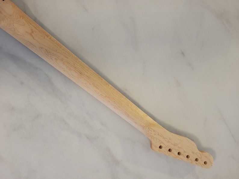 May include: A light-colored, unfinished guitar neck with six tuning peg holes. The wood grain is visible, and the neck is angled diagonally against a light-colored background. This guitar part is ready for customization.