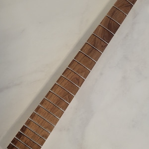 May include: A guitar neck with a light brown wooden fretboard and silver frets. The neck is unfinished and ready for installation. The headstock is a lighter color wood. The neck is angled diagonally across a white surface.