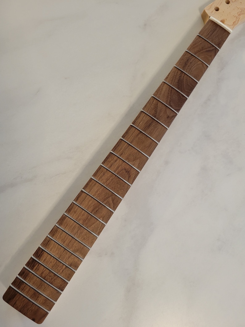 May include: A guitar neck with a light brown wooden fretboard and silver frets. The neck is unfinished and ready for installation. The headstock is a lighter color wood. The neck is angled diagonally across a white surface.