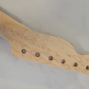 May include: A light-colored, unfinished guitar neck with a headstock featuring six circular holes for tuning pegs. The wood grain is visible, and the neck is angled slightly to the left. The guitar neck is a musical instrument part.