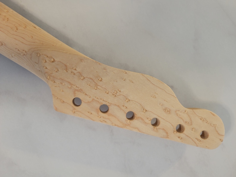 May include: A light-colored, unfinished guitar neck with a headstock featuring six circular holes for tuning pegs. The wood grain is visible, and the neck is angled slightly to the left. The guitar neck is a musical instrument part.