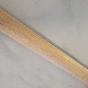 May include: Close-up of a light-colored wooden guitar neck. The wood grain is visible, with a pattern of small, dark spots. The neck is angled, showing the back and side.