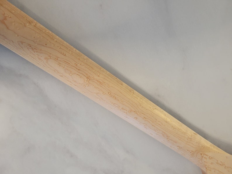 May include: Close-up of a light-colored wooden guitar neck. The wood grain is visible, with a pattern of small, dark spots. The neck is angled, showing the back and side.