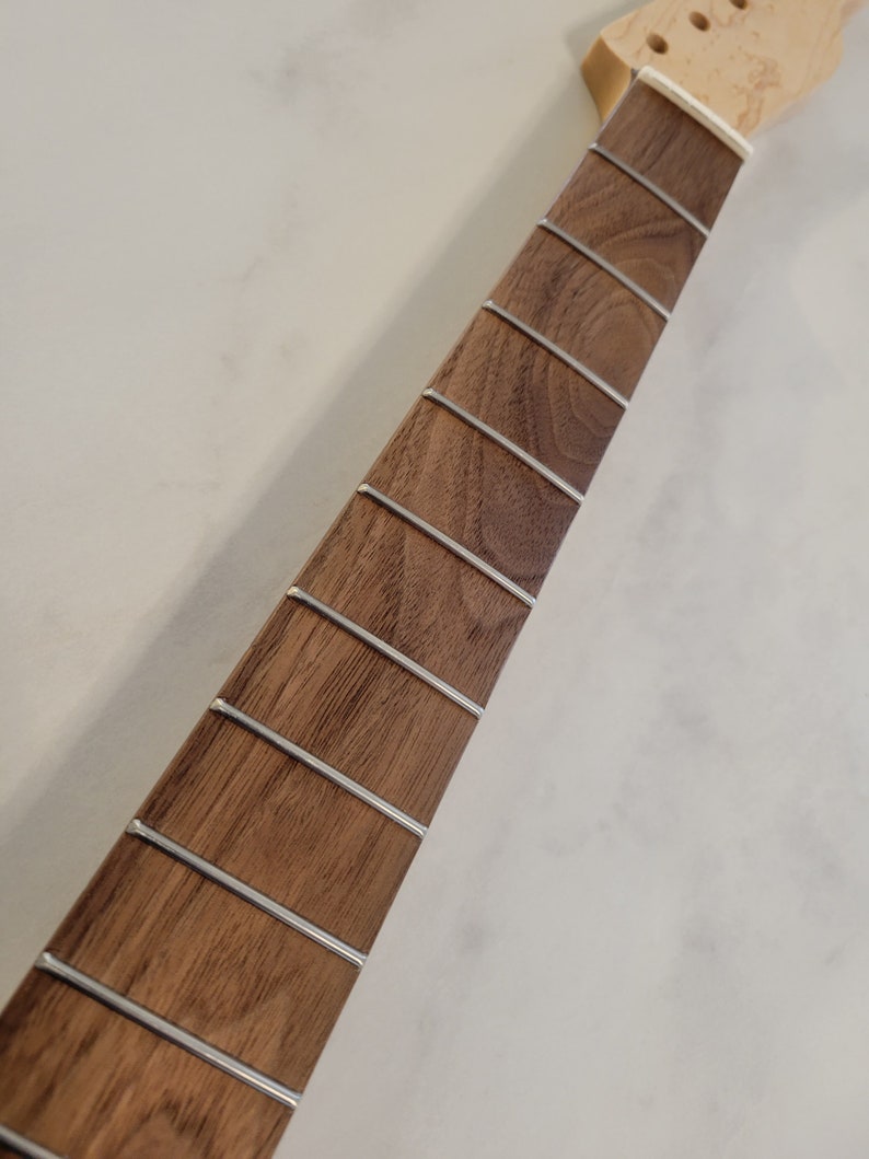 May include: Close-up of a guitar neck with a dark brown fretboard and silver frets. The headstock is unfinished, showing the natural wood grain. The neck is a musical instrument component.