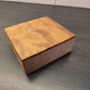 Peut inclure: Une petite boîte carrée en bois avec un couvercle brun clair et une base brun plus foncé. Le couvercle présente un motif de grain de bois naturel. La boîte est probablement utilisée pour ranger ou exposer de petits objets. La boîte est posée sur une surface gris foncé.