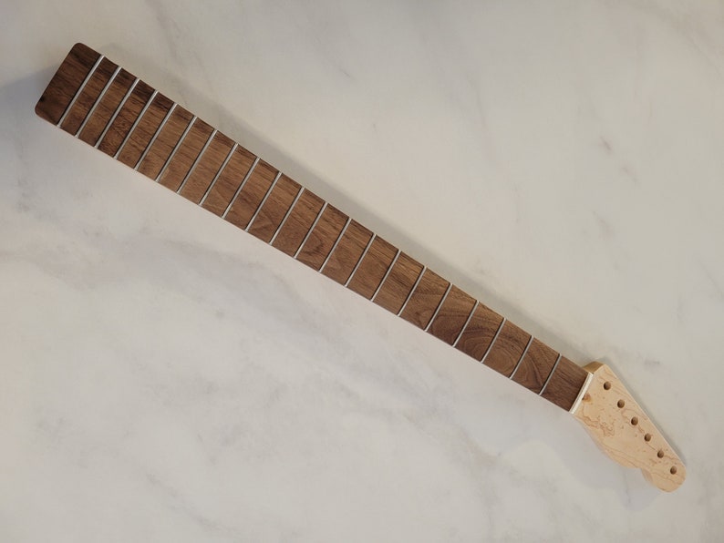 May include: A guitar neck with a dark brown fretboard and silver frets. The headstock is unfinished light wood with pre-drilled holes for tuning pegs. The neck is angled on a white surface.
