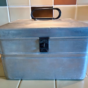 May include: A rectangular silver-colored metal lunch box with a matte finish. The container features a hinged lid, a black plastic oval-shaped handle on top, and a central black metal latch mechanism on the front for securing the lid closed.