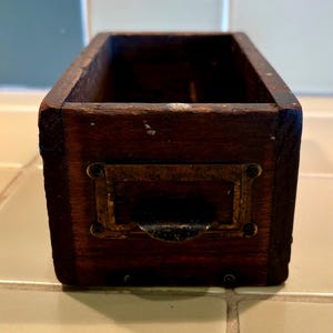 May include: A rectangular, dark brown wooden storage box featuring a weathered finish and a metal label holder attached to the front. The box has an open top and a curved, dark metal pull handle integrated into the brass-toned label frame.