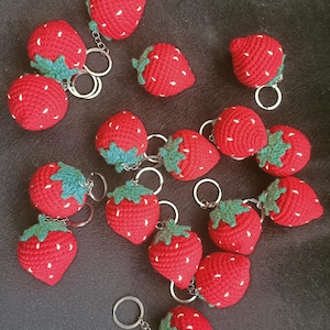 Puede incluir: Una colección de 16 llaveros de fresa hechos a crochet. Cada llavero es rojo con hojas verdes y semillas blancas. Los llaveros están dispuestos sobre una superficie gris oscura.