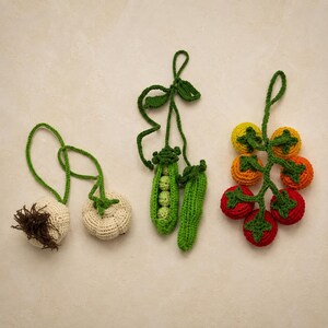 May include: Three sets of crocheted vegetable ornaments featuring a pair of off-white garlic bulbs with brown roots, a green pea pod with visible round peas inside, and a cluster of six small tomatoes in shades of red, orange, and yellow.