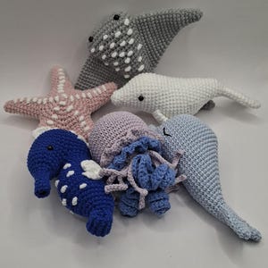 Könnte beinhalten: Eine Sammlung handgefertigter gehäkelter Meeresbewohner, darunter ein grauer Stechrochen mit weißen Punkten, ein rosa Seestern, ein weißer Delfin, ein blaues Seepferdchen, eine lavendelfarbene Qualle und ein hellblaues Narwal. Ideal für Meeresdeko.