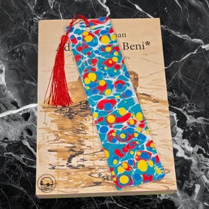 May include: A colorful bookmark with a marbled design in blue, red, and yellow, resting on a book. The bookmark has a red tassel attached to the top. The book has text on the cover.