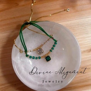 Peut inclure: Deux bracelets superposés avec des perles vertes et des accents dorés. Les bracelets présentent une pierre précieuse verte carrée et une pierre précieuse transparente carrée. Les bracelets sont présentés dans un bol blanc avec le texte "Divine Alignment Jewelry".