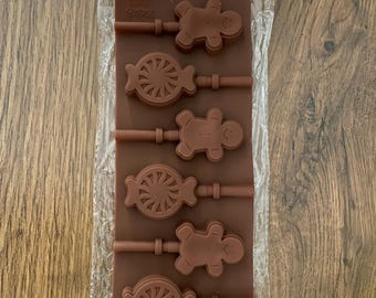May include: A brown silicone mold for making lollipops, featuring three gingerbread man shapes and three circular swirl patterns. The mold includes integrated channels for sticks and comes with a set of white plastic sticks in a clear plastic bag.