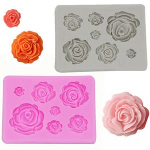 May include: Two silicone moulds for making rose-shaped decorations. One mould is grey and has seven rose shapes. The other mould is pink and has eight rose shapes. There are three finished rose decorations in the image, one orange, one pink, and one red.