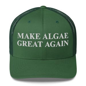 Peut inclure: Une casquette de type trucker vert forêt avec un panneau arrière en filet et un panneau avant solide. Le texte "MAKE ALGAE GREAT AGAIN" est brodé en lettres capitales blanches au centre du panneau avant. La casquette a une visière courbée.