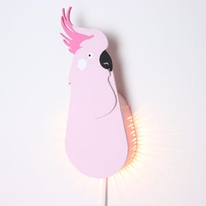 May include: A pink wooden wall lamp shaped like a cockatoo with a black eye and beak. The lamp has a warm white light that shines through the cutouts in the back of the lamp.