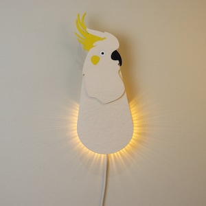 May include: A white cockatoo-shaped wall lamp with yellow crest feathers and a black beak. The lamp is illuminated, casting a warm glow on the wall.