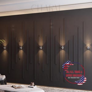 Op de afbeelding: Een donkergrijze muur met verticale, verhoogde rechthoekige accenten en verschillende kleine, ronde, zilverkleurige wandkandelaars. De wandkandelaars geven een warm licht af. De afbeelding bevat ook een kleine tafel en een decoratief element.