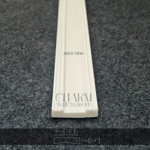 K&ouml;nnte beinhalten: Wei&szlig;e Polyurethan-Leiste mit dekorativem Design. Die Leiste ist 1,6 Zoll breit und 0,4 Zoll dick. Die R&uuml;ckseite der Leiste ist im Bild zu sehen. Der Text "CHARM wallcreations" ist auf die Leiste gedruckt.
