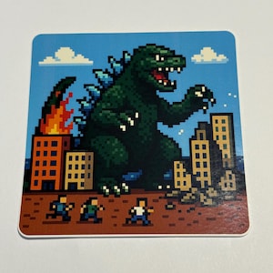 May include: Square coaster featuring a pixelated Godzilla destroying a city. The green monster is depicted with a menacing expression, towering over buildings. Flames erupt from a building, and tiny figures flee the scene. The background is blue with white clouds.