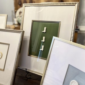 Puede incluir: Obra de arte enmarcada con tres pequeñas botas de vaquero blancas sobre un fondo verde. Las botas están dispuestas verticalmente dentro de un marco dorado, sobre un passe-partout blanco. Se ven otras piezas enmarcadas, con marcos dorados similares.