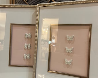 May include: Two rectangular wall frames with gold-toned textured borders and off-white matting. One frame contains three small white bows arranged vertically, while the other frame features three small white butterflies in a vertical alignment.