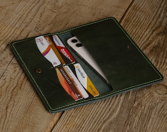 May include: Open, dark green leather wallet with a snap closure. The wallet is open, revealing a smartphone and several credit cards. The stitching is a lighter green, and the wallet is on a wooden surface.