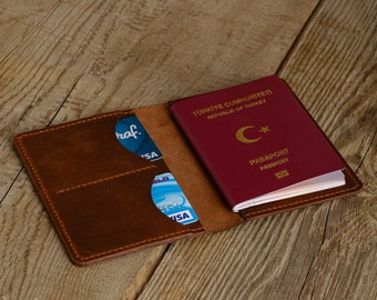 May include: A brown leather passport holder with a red Turkish passport inside. The passport holder has two slots for cards and a pocket for cash. The passport holder is open and the cards are visible.